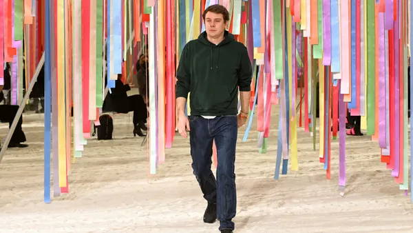 A person in a black hooded sweatshirt and jeans walks down a runway. In the background are multicolored ribbons hanging from the ceiling.