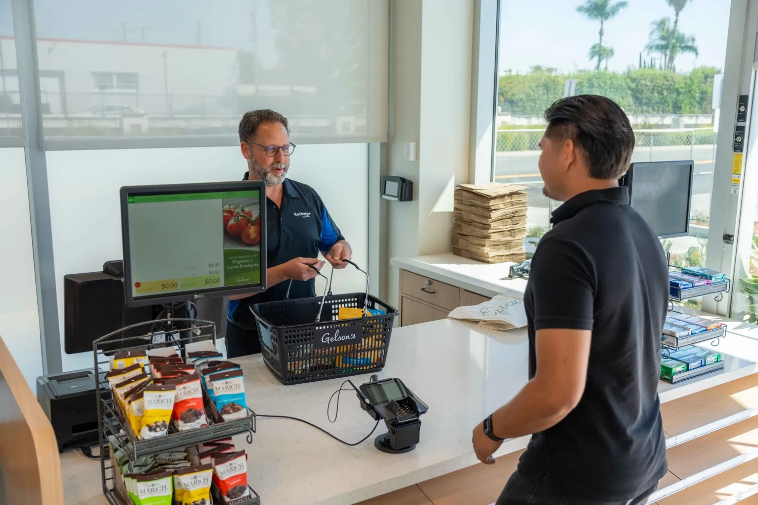 A photo of a worker standing behind the counter at a checkout location at a store. Another person stands on the other side of the counter. There is a cash register on the counter, and the outdoors can be seen through a window behind the worker.