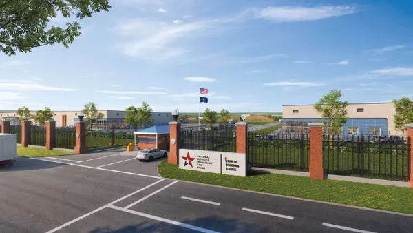 A black fence connected by brick, with a security checkpoint at the building entrance and two white buildings in the background.