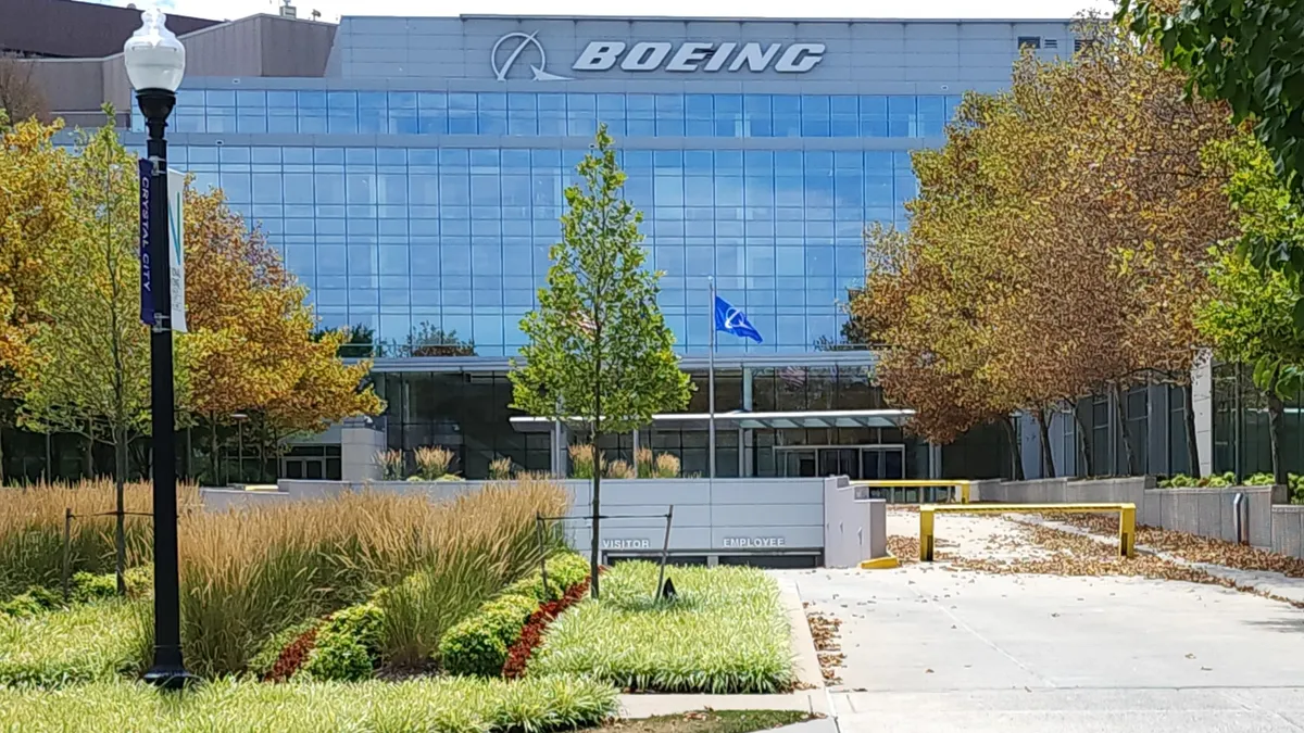 A building with glass windows and the white Boeing logo in between rows of trees and green landscape in front of it.