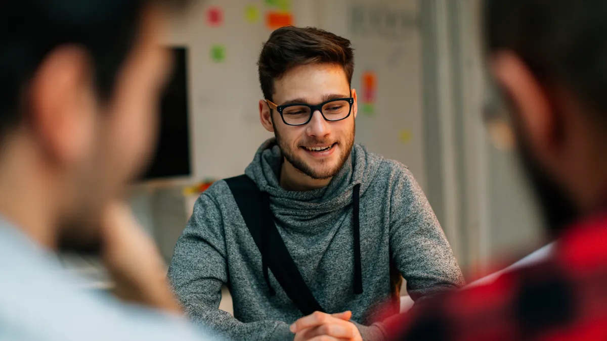 Young man at a job interview talking with two developers. They are running startup developer company from their home. Over the shoulder view of them discussing.