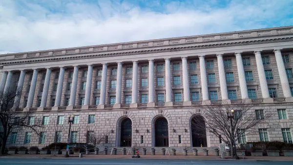 A low-angle, wide-shot image of a building depicting Classical Revival architecture, featuring a symmetrical façade with tall, evenly spaced windows and prominent stone columns, is set in front of a blue sky.