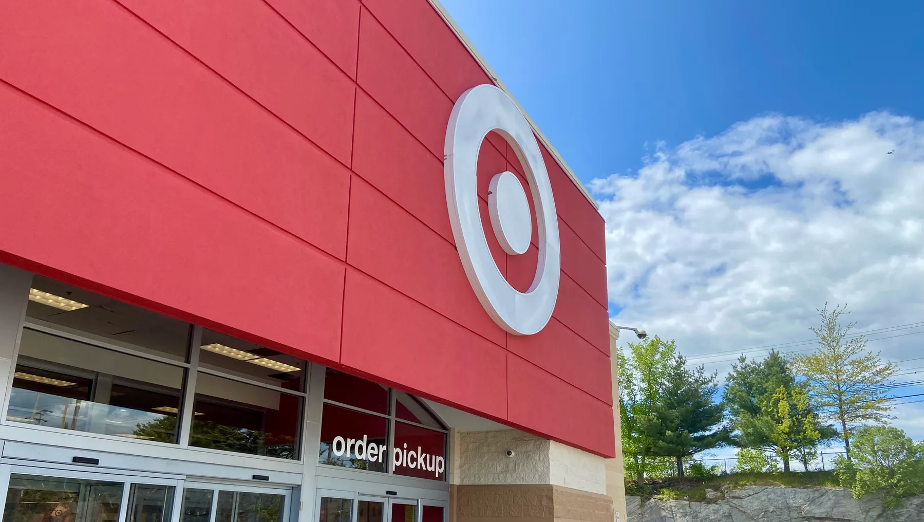 A red storefront entrance with a white dot-in-circle logo and an "order pickup" sign on the door. Trees and blue sky with white clouds on the right.