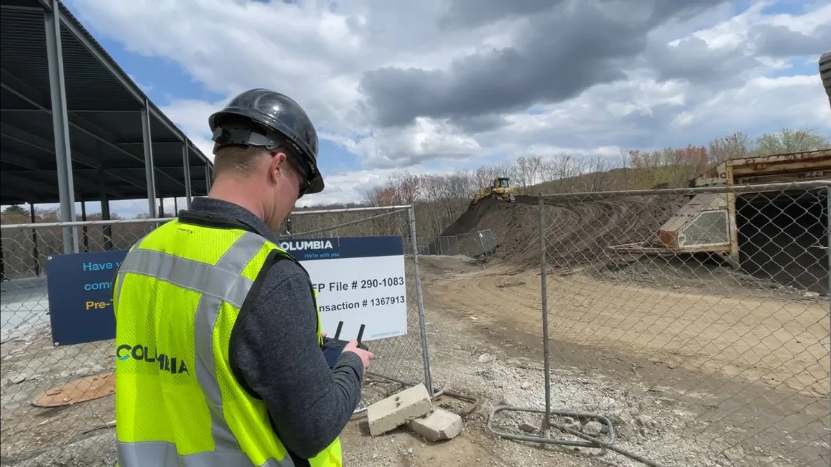 A person in a yellow safety vest and hardhat holds a remote in their hand on a construction site.