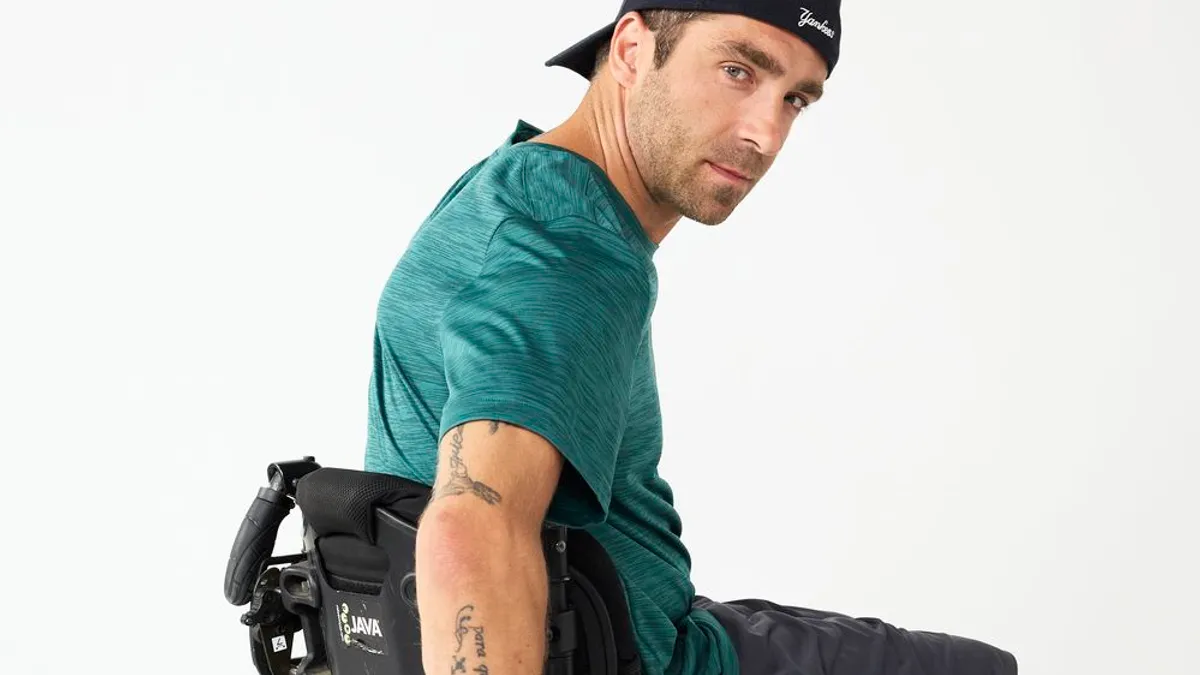 A person in a wheelchair, wearing a black baseball-style cap, gives an over-the-shoulder glance, hands gripped on the chair's back wheels.