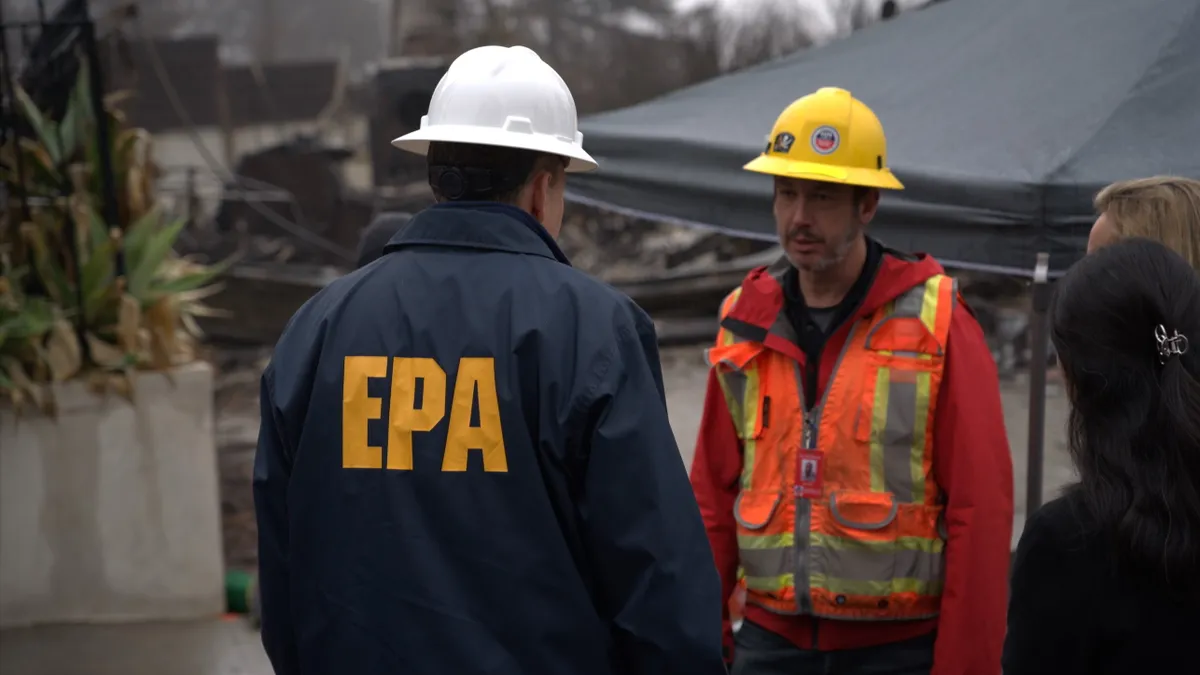 A person wearing a jacket with "EPA" emblazoned on the back meets outside a tent with a worker wearing high-visibility clothes and a helmet.