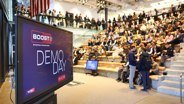 A group of people sit in an auditorium while a screen shows BOOST Demo Day in the foreground.