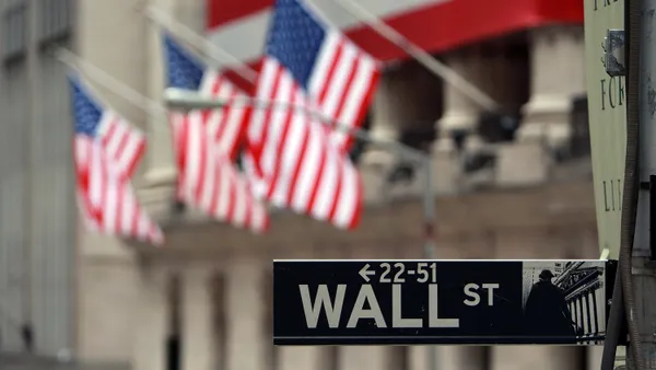 The Wall Street street sign is visible in the foreground with a picture of a trader in front of the stock market on the left of the sign. Three american flags are out of focus in the background.