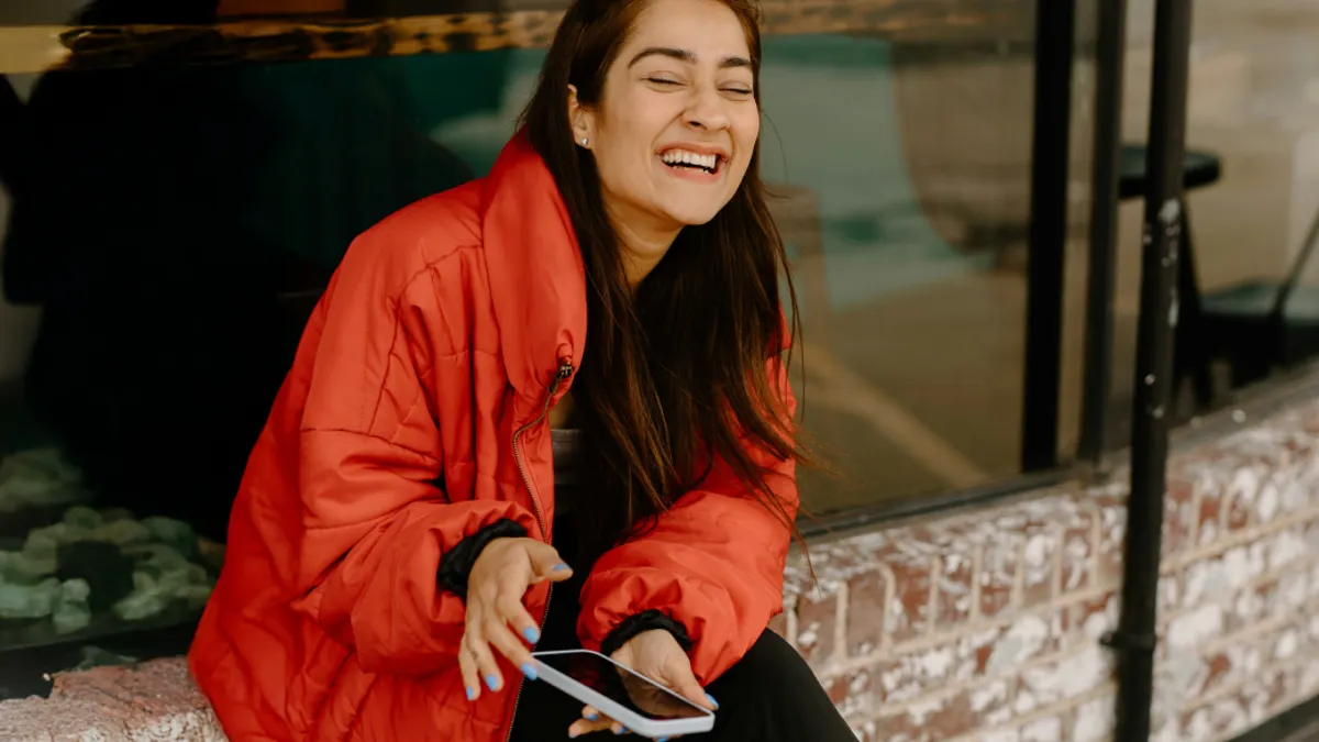 A woman in a red jacket sits on a brick ledge, laughing joyfully while holding a smartphone. Her expression conveys happiness and warmth.