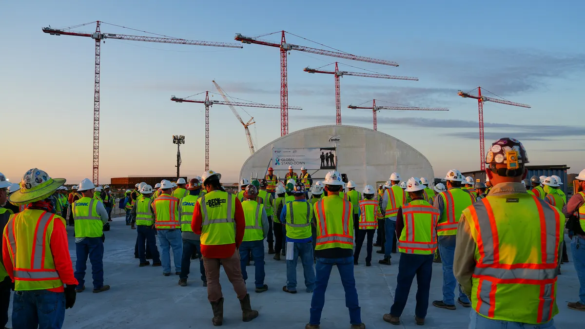 A large group of construction workers stand around for a meeting before a backdrop of towering cranes.