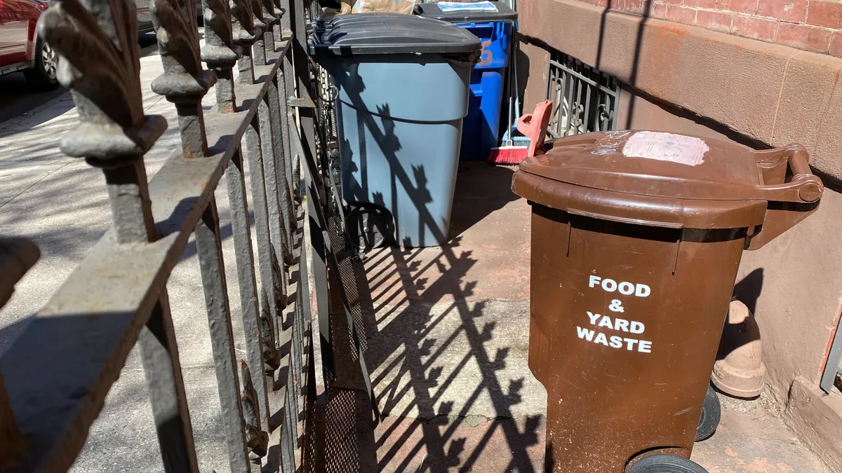 Several large plastic waste bins sit directly in front of a building in a fenced-off area. One of them reads "Food & Yard Waste"