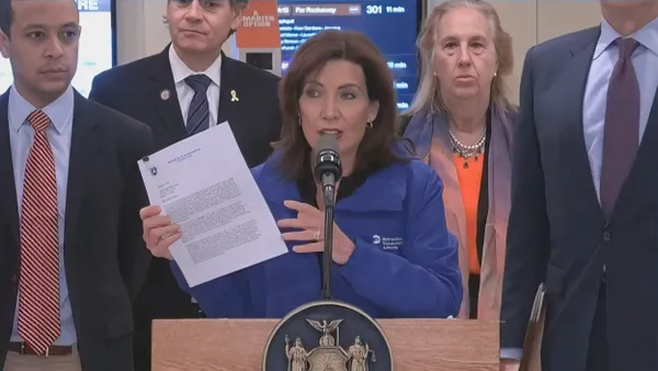 A woman in a blue jacket at a podium holding up a letter surrounded by several people.