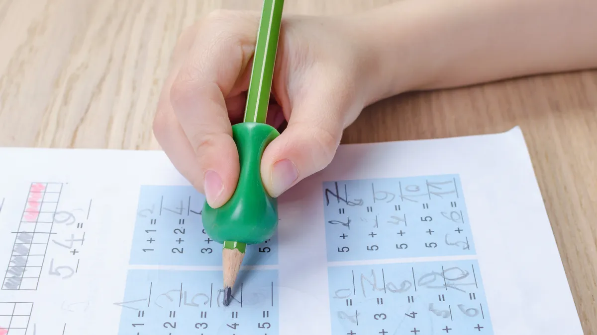 A close-up of a student holding a pencil with a finger grip. The pencil is pointed at a paper on a desk.