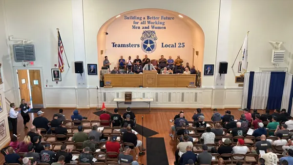 A view looking down at a union hall from a balcony. Behind the stage on the wall are the words "Teamsters Local 25" and the IBT logo. People are gathered on stage and seated in the audience.