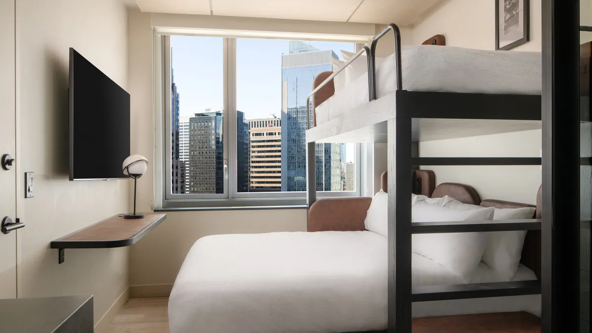 Bunk beds with white bedding sit before a window with a view of Manhattan skyscrapers.