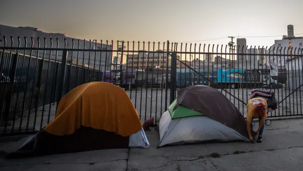 A person secures one of two tents set up in front of a tall gate with empty lot and buildings in the background.
