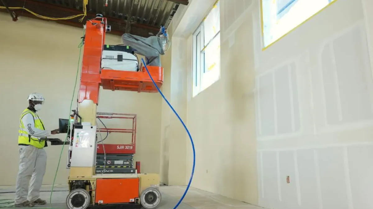 A worker in safety gear looks on as a robot works on a wall. The robot is tall and orange.