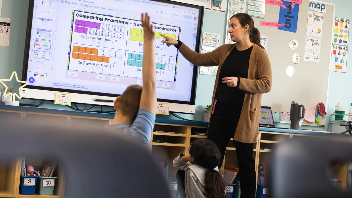 An adult stands in front of a classroom and points to a large screen on a wall. The screen has information about comparing fractions.