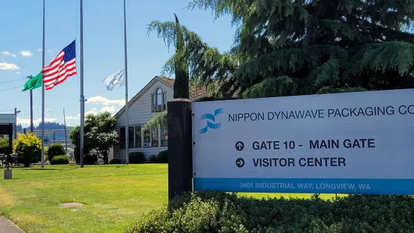 Flags fly at half staff outside an industrial facility with a sign that says Nippon Dynawave Packaging Co.