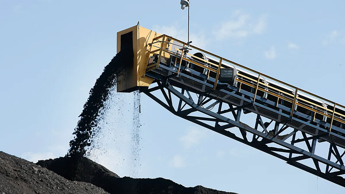 Coal falls off a conveyor belt onto a larger pile of coal. Multiple mounds of coal can be seen.