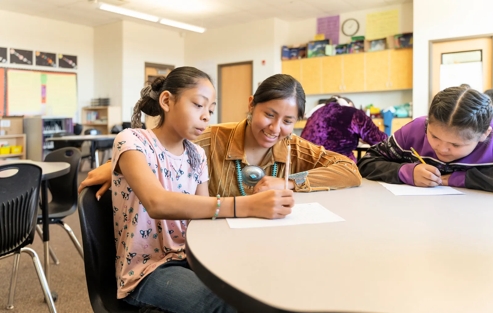 An adult is with two students at a desk in a classroom. The students are writing and reading.