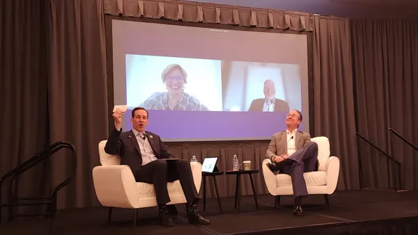 Two people in suits sitting on chairs on stage, and two people on the screen behind them.