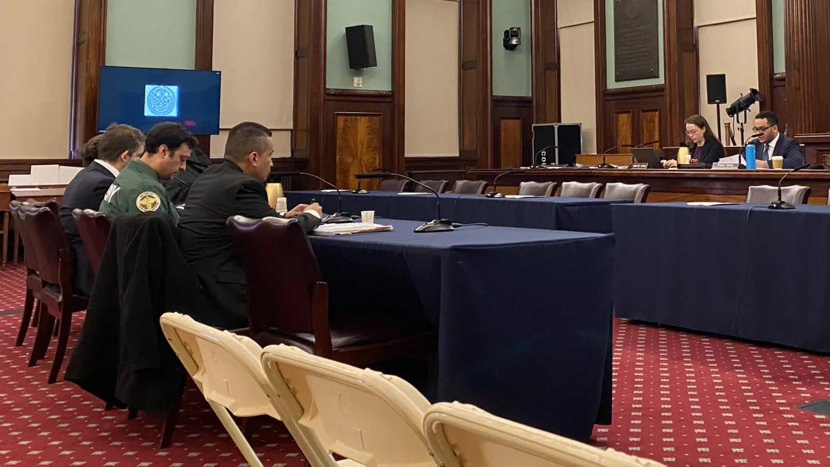 A council chamber where a man and woman in suit sit on dais while four people, one wearing a jacket labeled DSNY, sit at a table facing the dais.