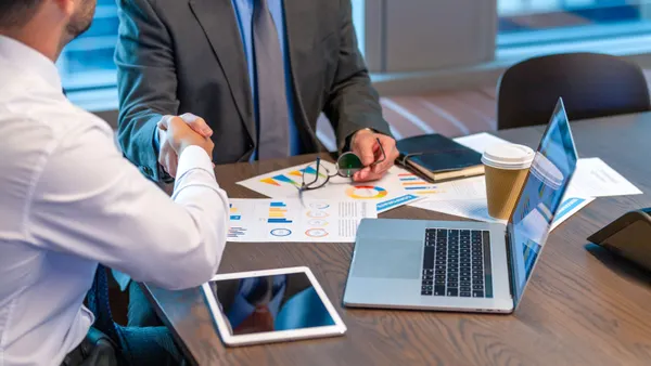 Businessmen shaking hands. Both are dressed in business clothing and there is paperwork with finance graphs and charts. There is also technology including a laptop computer and digital tablet