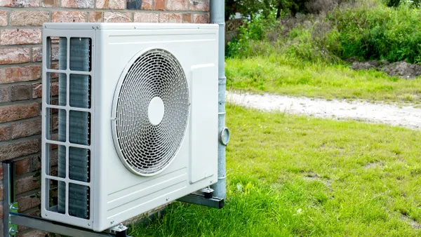 A white box with a circular fan grill sits in front of a brick house.