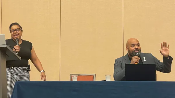 Two people are on a stage and holding microphones. One person is standing and one person is seated.