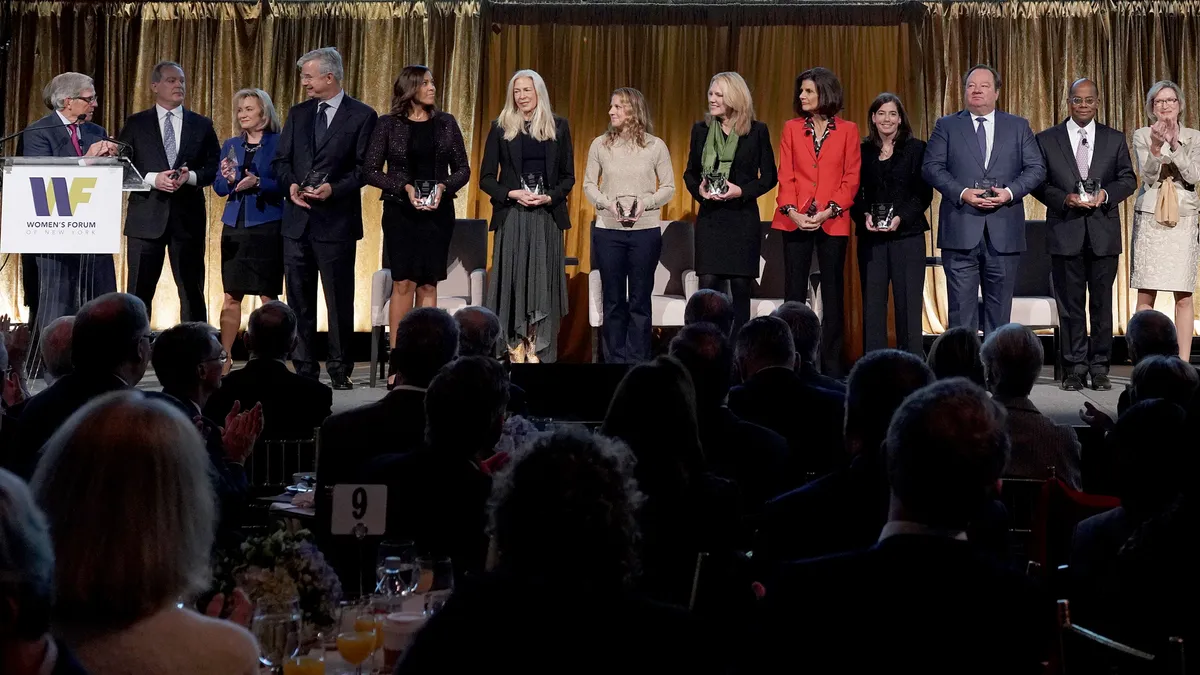 A row of diverse people stands on stage with a podium to the left of the stage and in front of a clapping crowd.