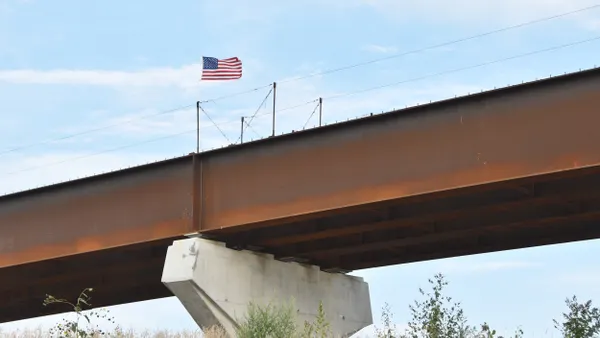 A U.S. flag flies over rusty beams on a concrete bridge under construction.