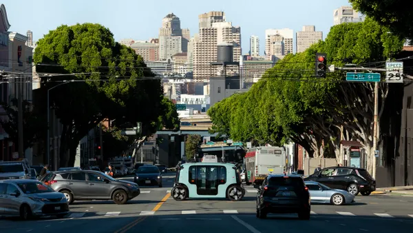 A light blue shuttle vehicle crosses a busy intersection against the San Francisco skyline.