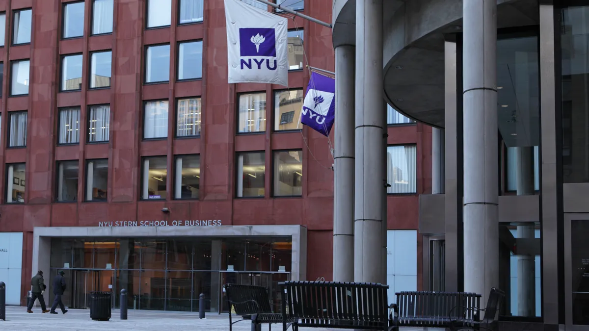 Two unknown men walk into a red and glass paneled building that says NYU Stern School of Business with NYU flags flying on the right side of the photo.