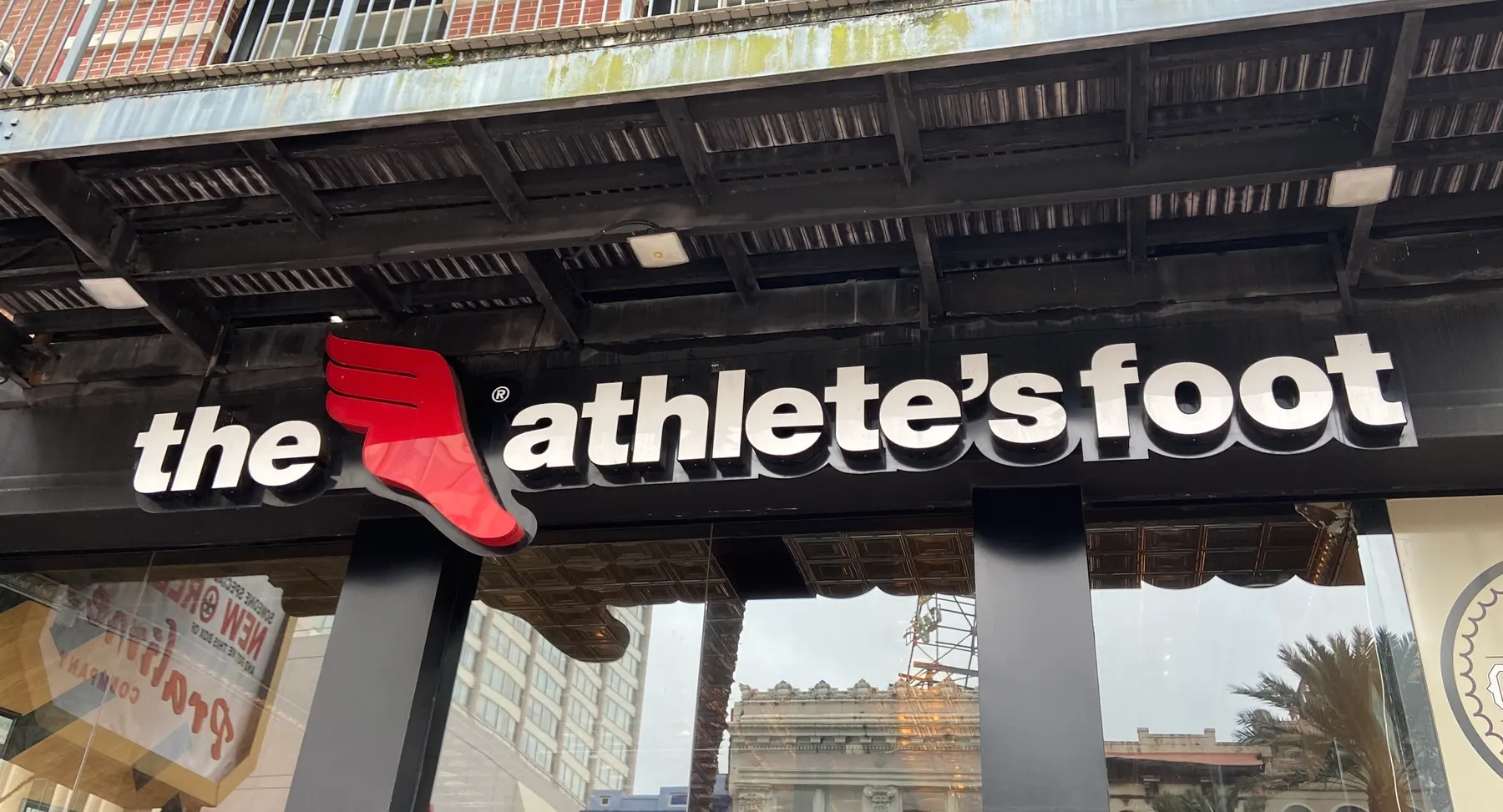 Buildings and signs are reflected in a store window. Above a sign in white letters reads "the athlete's foot," with a red symbol of a winged foot.