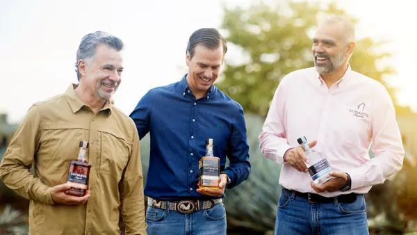 Three men in different colored button-up shirts each hold a bottle of Lunazul as they walk through a field.