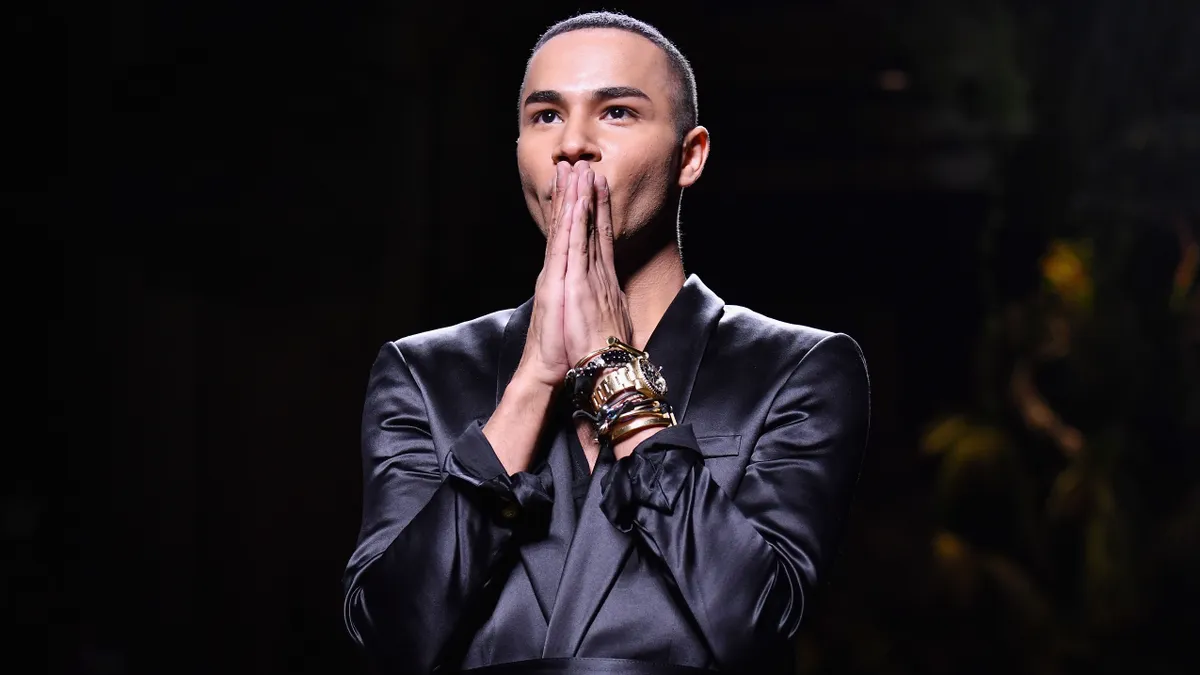 Balmain creative director Olivier Rousteing stands in a black satin jacket and clasps his hands in front of his face in a gesture of gratitude.