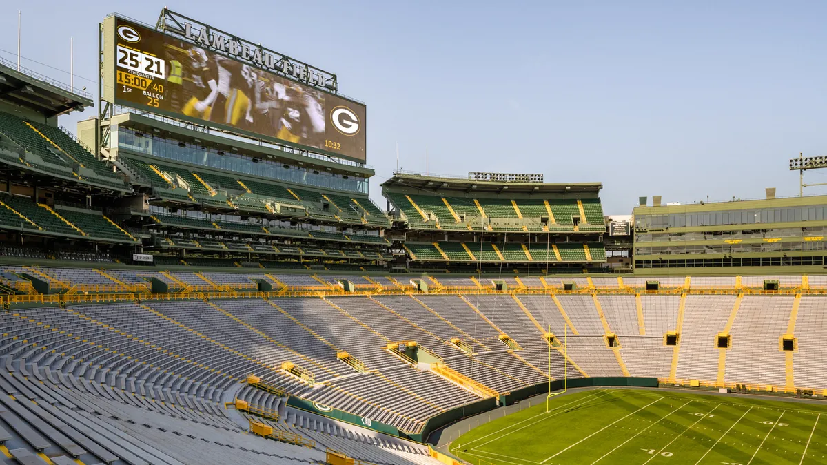 A shot from the stands at Lambeau Field. Stands and seating surround the grass field below, and a scoreboard above plays an old video.