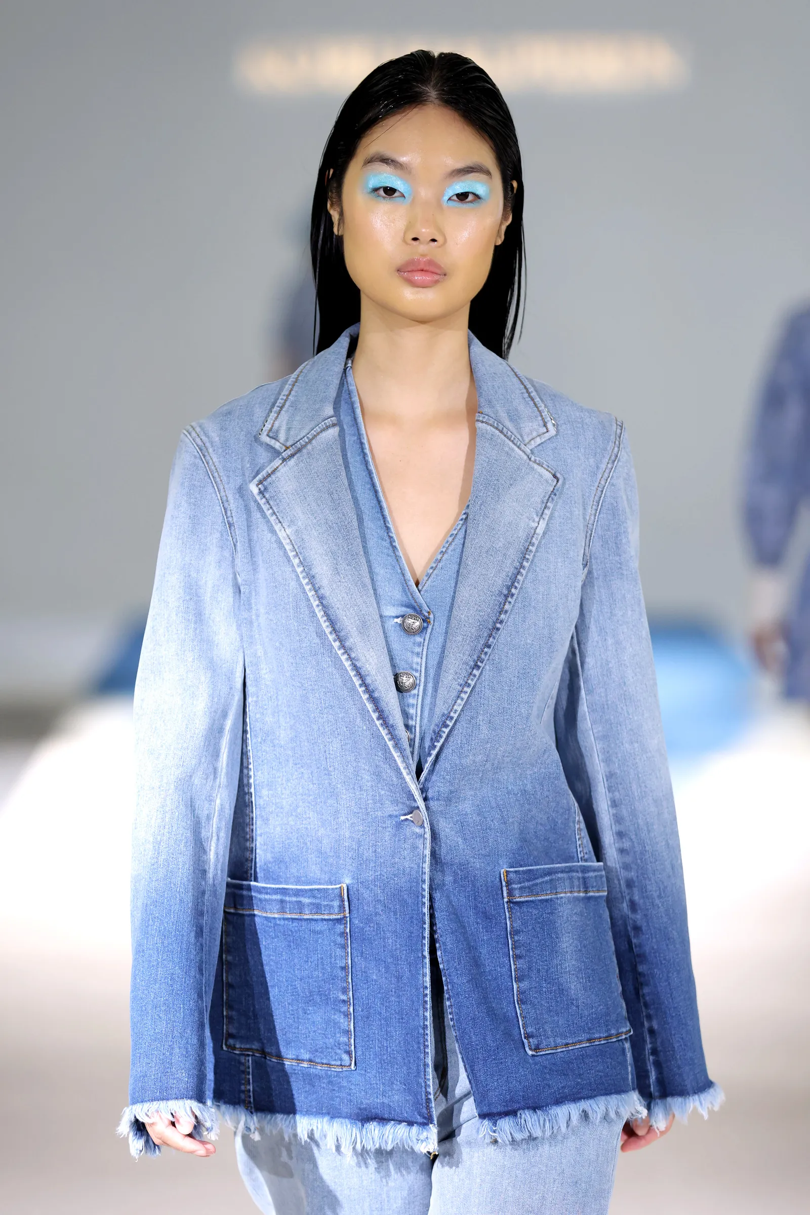 A model in blue eyeshadow and a denim suit walks down the runway.