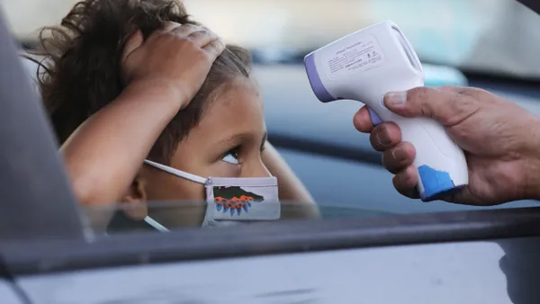 A young student is wearing a face mask and sitting in a car. The car window is down and the child has pushed their hair on their forehard back. Another person's arm holds a thermostat to the child's forehead.