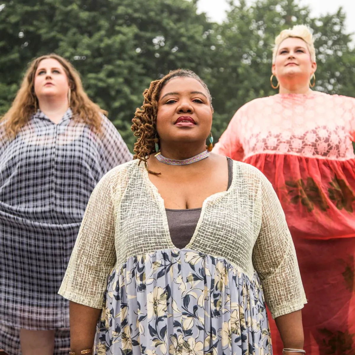 Three women stand outside during the day wearing dresses. A line of trees is in the background.