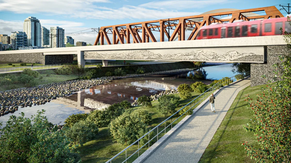 A red train passes on an open bridge spanning a stream and walking path.