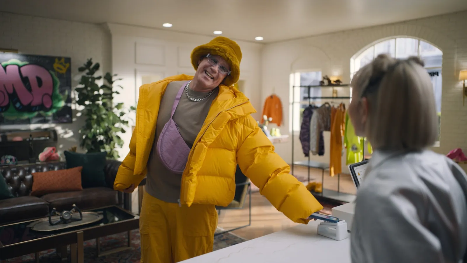 Actor Will Ferrell stands in retail boutique wearing a yellow outfit  with arm outstretched using a phone to pay in front of an sales counter.