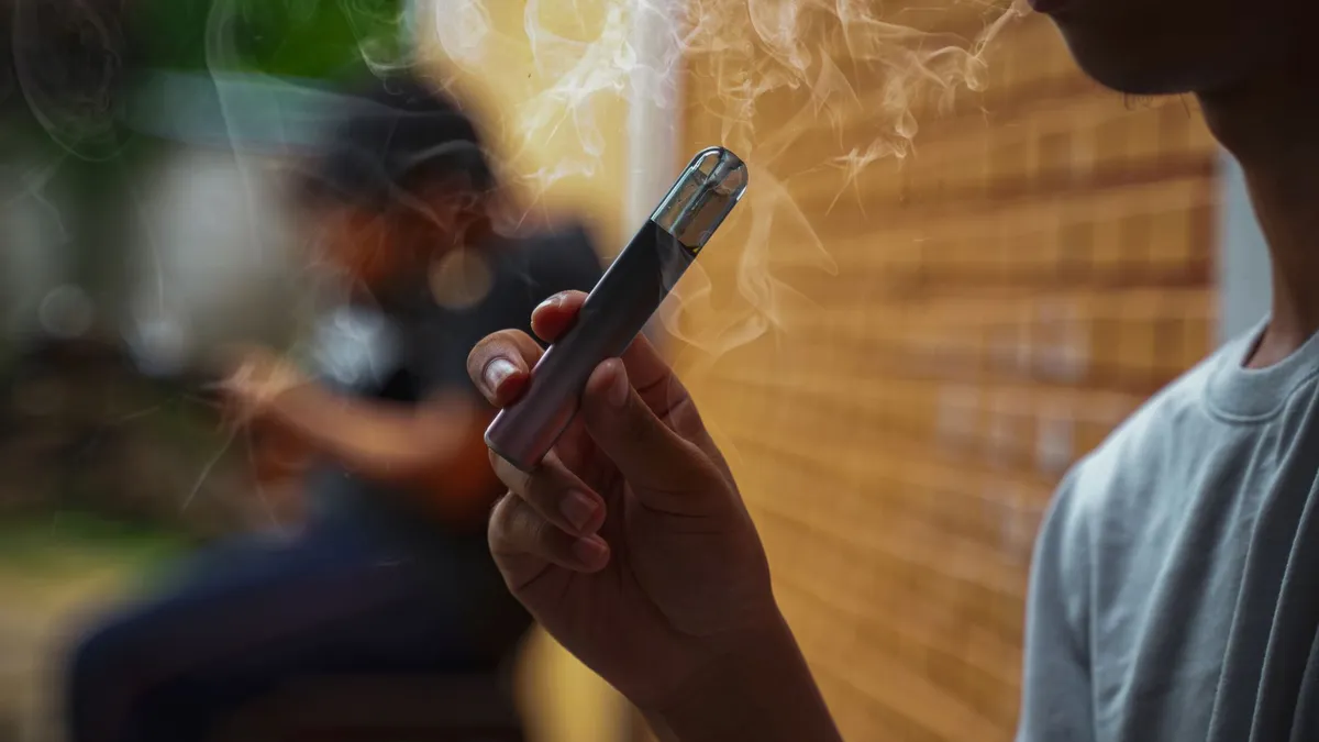 Male teenager holds a smoked e-cigarette in his hand while sitting during recess in a hidden corner with his friends at school