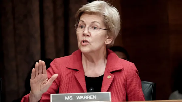 Senator Elizabeth Warren holds out her hand as she speaks. In front of her a placard reads "Ms. Warren."