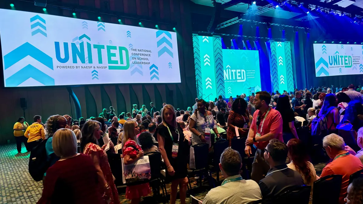 People are sitting or standing in a large room with lighted screens in the front that read "United"