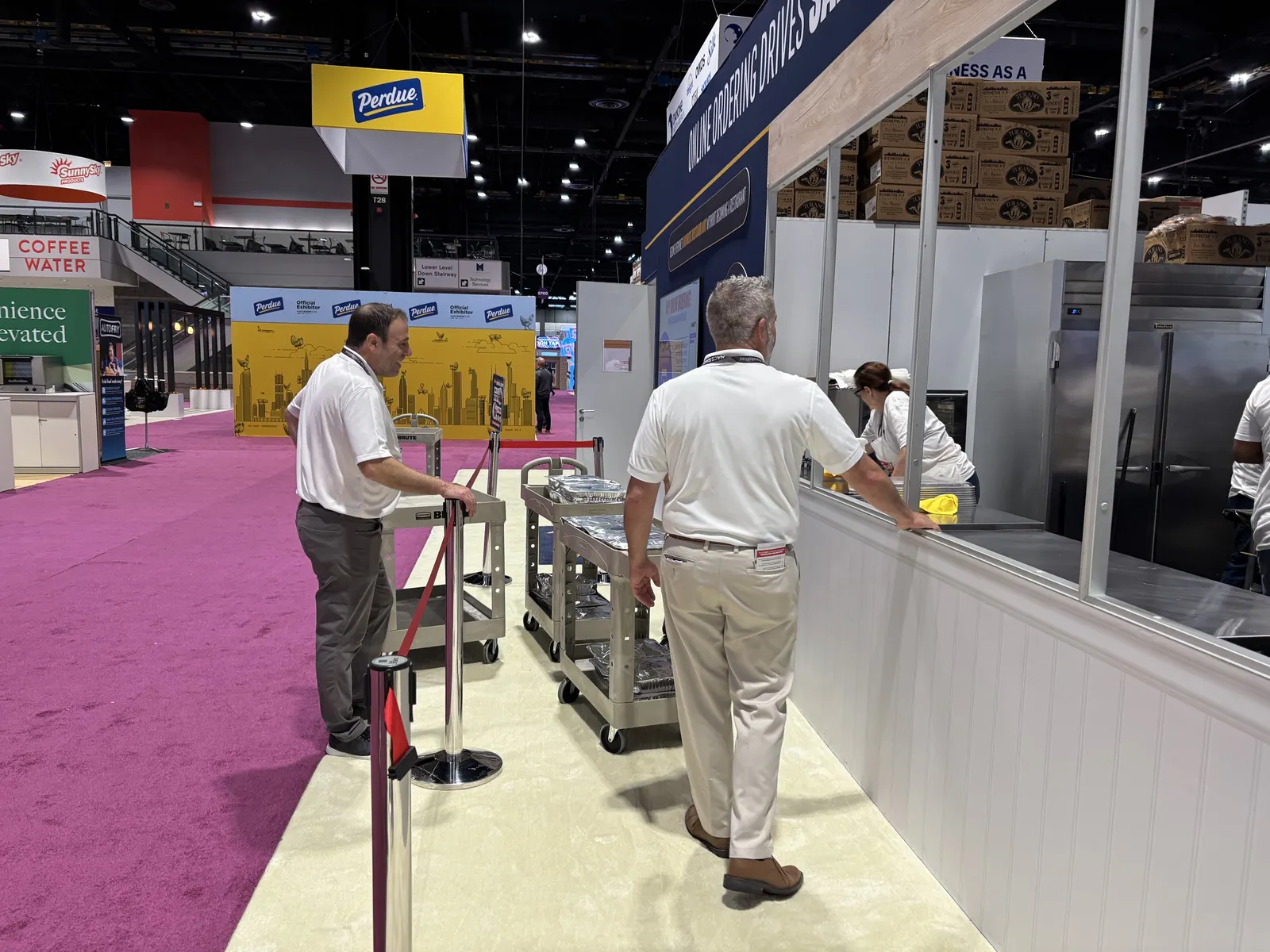 Several people stand around trolleys holding metal pans of food items in an expo center.