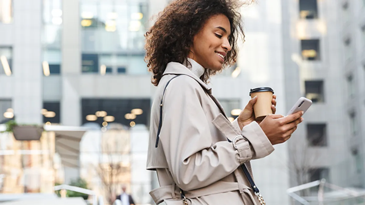 Smiling woman wearing coat walking outdoors, using mobile phone