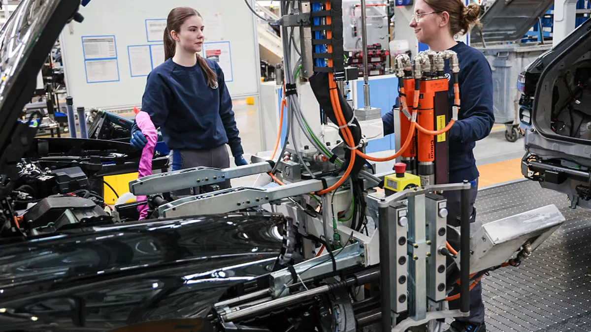 Two women work complex machinery to aid in the building of a car.