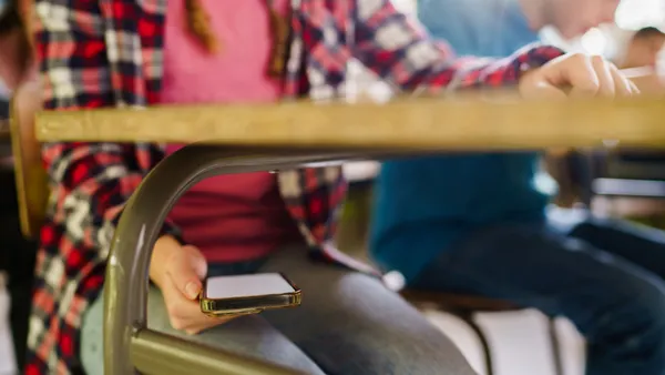 A student is pictured sitting at a desk in a classroom holding a cellphone under the desk. Only the student's middle body is shown.